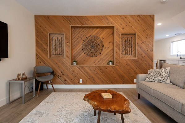 A modern living room with a wooden herringbone wall, a grey sofa, a brown leather ottoman, and a grey armchair.
