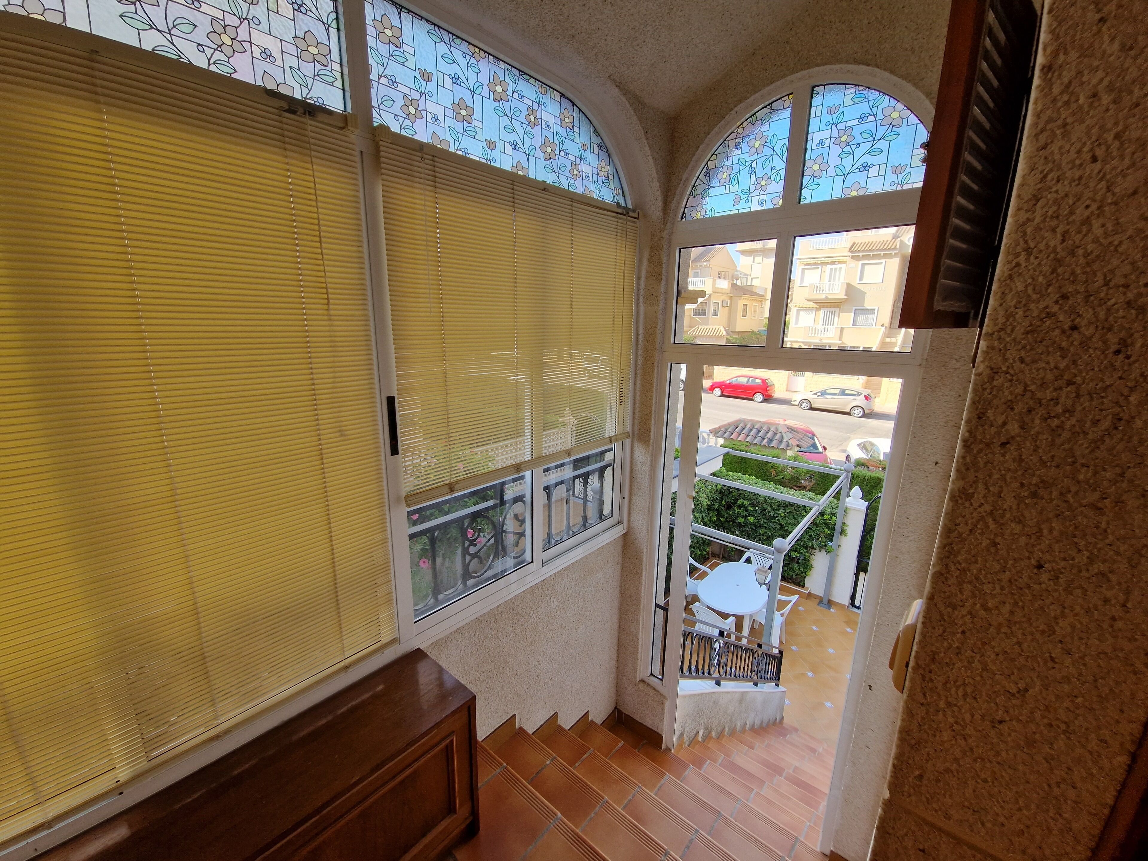 A room with a view of a street, a balcony with a table and chairs, and a window with blinds.
