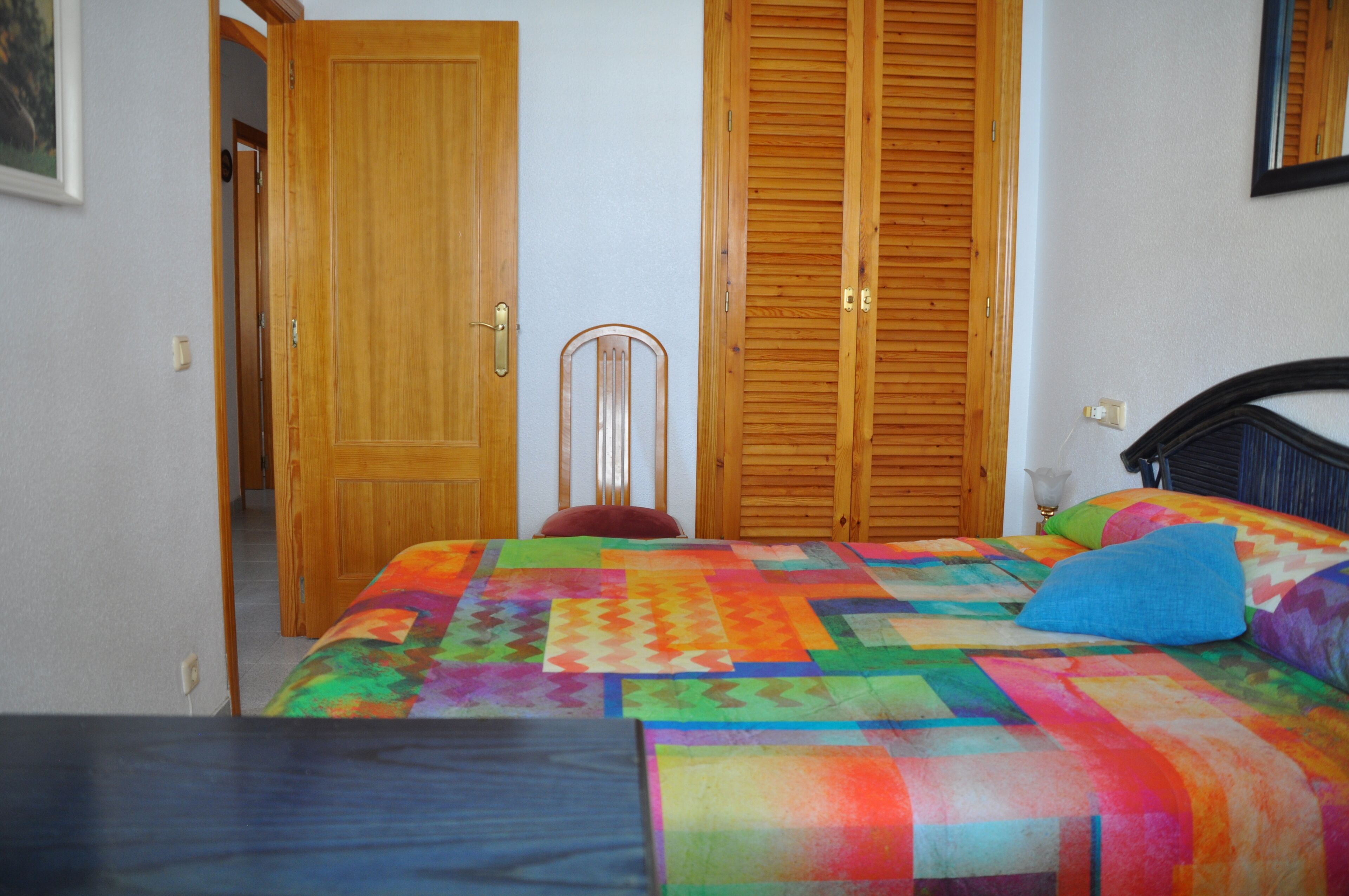 Ein Schlafzimmer mit farbenfroher Bettdecke, einem Holzschrank und einem Sessel.