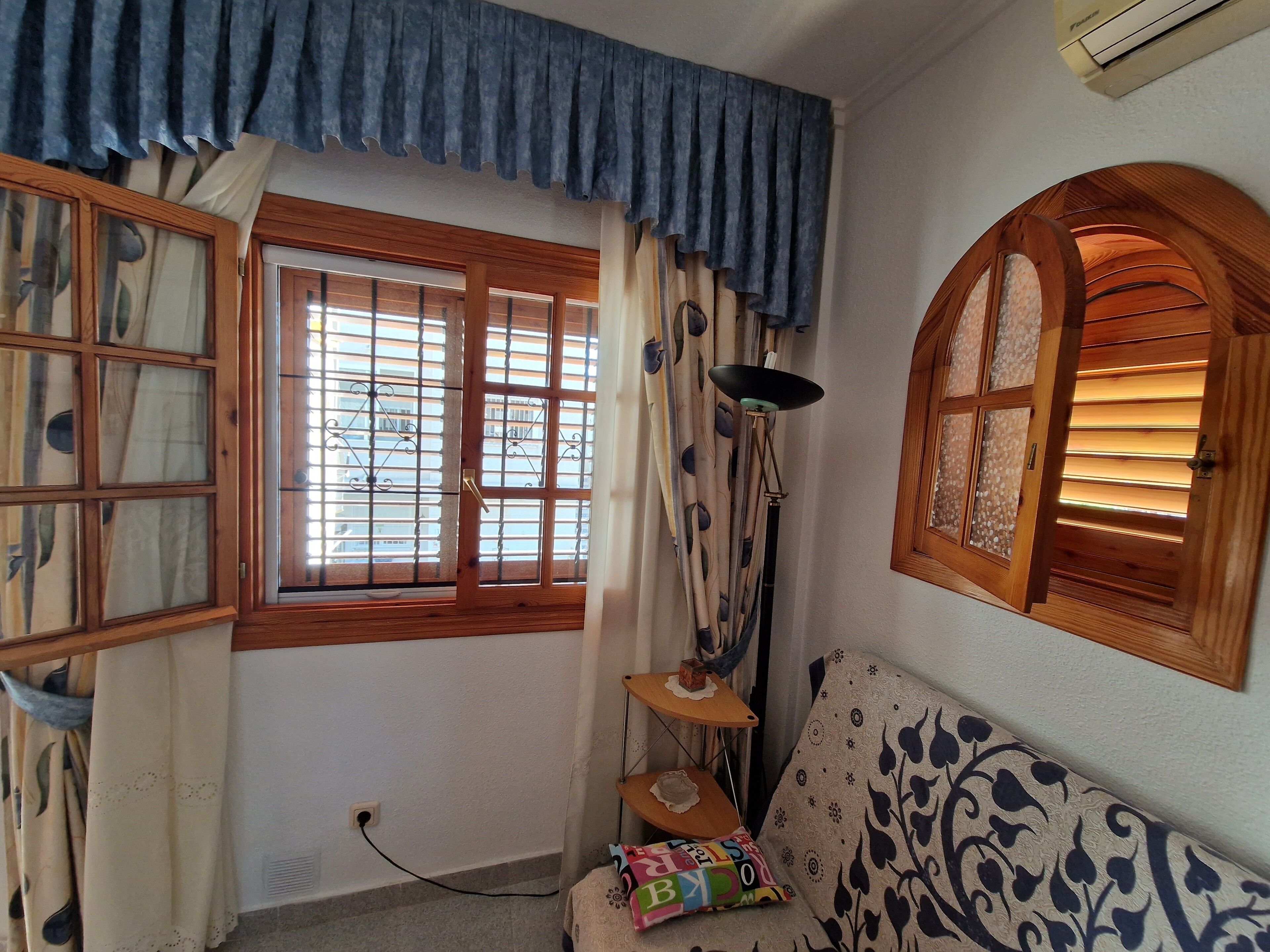 Quarto com uma grande janela com venezianas de madeira, um abajur, um sofá estampado e uma pequena mesa com luminária.