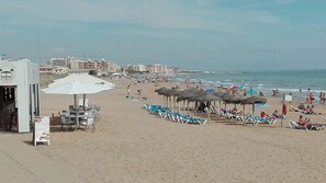 A beach with sun loungers and umbrellas, a building with a sign, and the ocean in the background. - Apartamento muy Luminoso / Playa de la Mata / Wifi / Patio / Terraza / Piscina (Torrevieja)