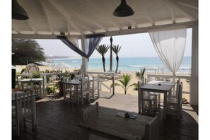 An outdoor seating area with white furniture, overlooking a beach with palm trees and the ocean. - Útulný apartmán in Praia de Chaves, Boa Vista (Sal Rei)