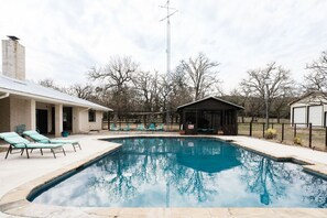 A swimming pool with lounge chairs, a covered seating area, and a brick building in the background. - Spacious Home on 5 Peaceful Acres | Pool & Game Room | Guest House (Fredericksburg)