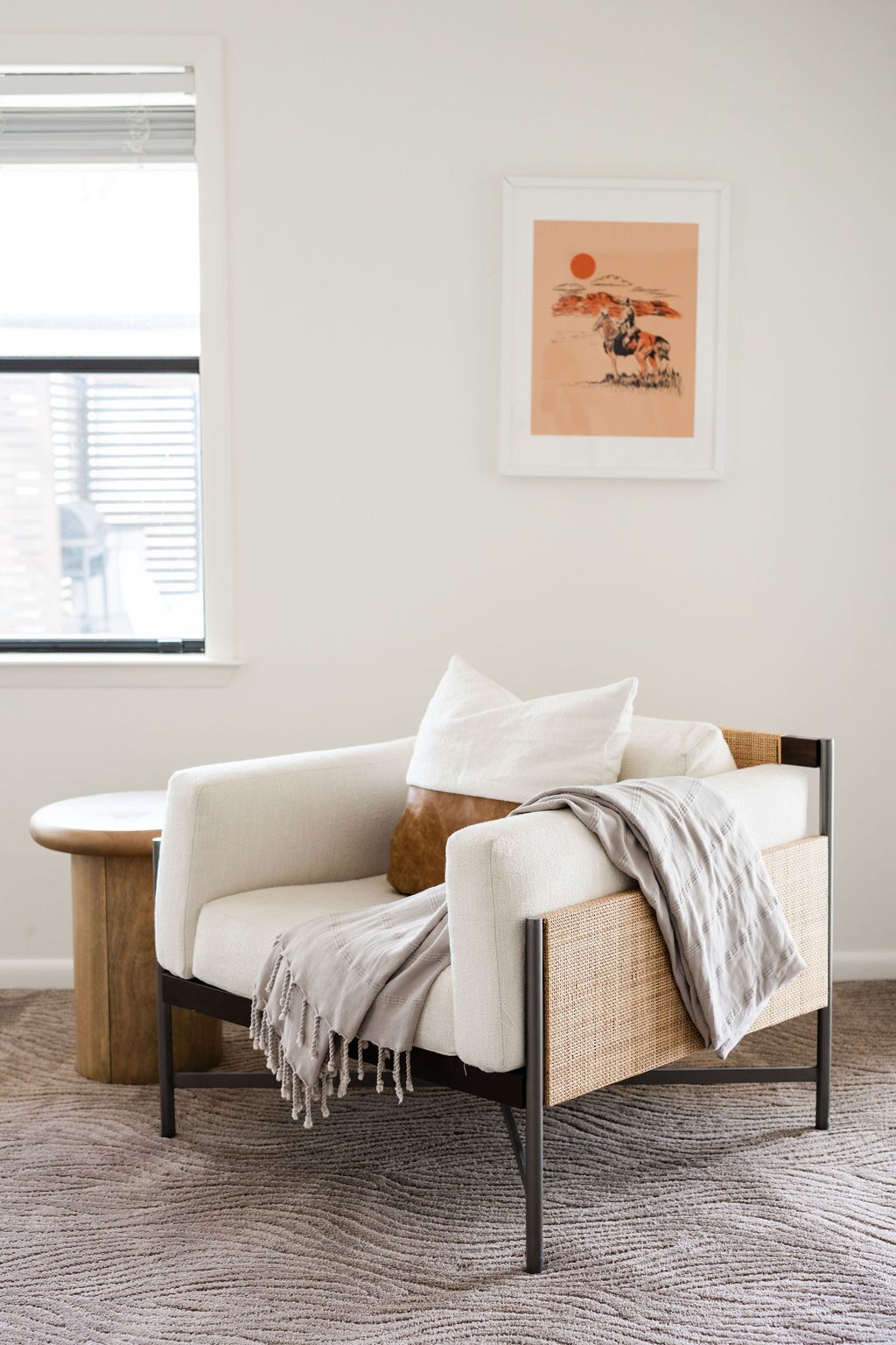 Ein modernes Wohnzimmer mit einem weißen Sofa, einem Holztisch und einem gewebten Sessel. An der Wand hängt ein gerahmtes Kunstwerk, das einen Sonnenuntergang mit einem Pferd zeigt.