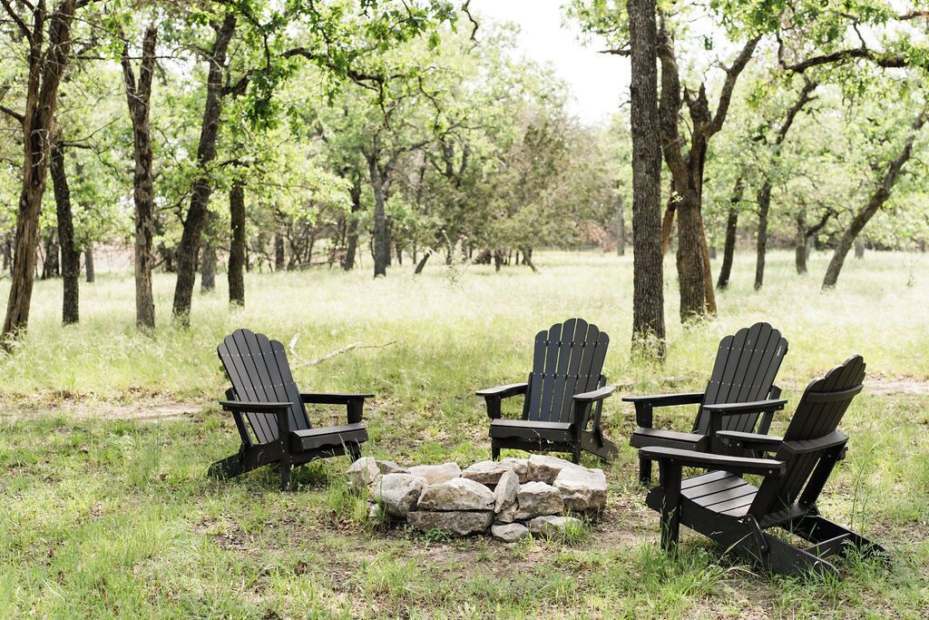 Tres sillas Adirondack dispuestas alrededor de un hogar de piedra en un espacio al aire libre con césped y árboles de fondo.