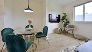 Un salon moderne avec une table à manger en verre, des fauteuils en velours vert, un luminaire suspendu noir, un téléviseur à écran plat, une plante en pot et une vue sur la mer par une grande fenêtre.