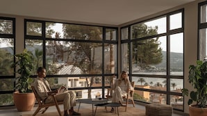 Amplia habitación con grandes ventanales que ofrecen vistas al mar y a los árboles. Hay dos sillas de madera y una pequeña mesa redonda.