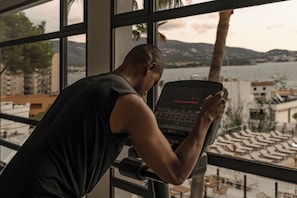 A person using a stationary bike with a view of a beach and palm trees through large windows. - Cook's Club Calvia Beach Adults Only +21 (Calvia)