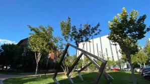Una escultura en un área verde con árboles y un edificio al fondo.