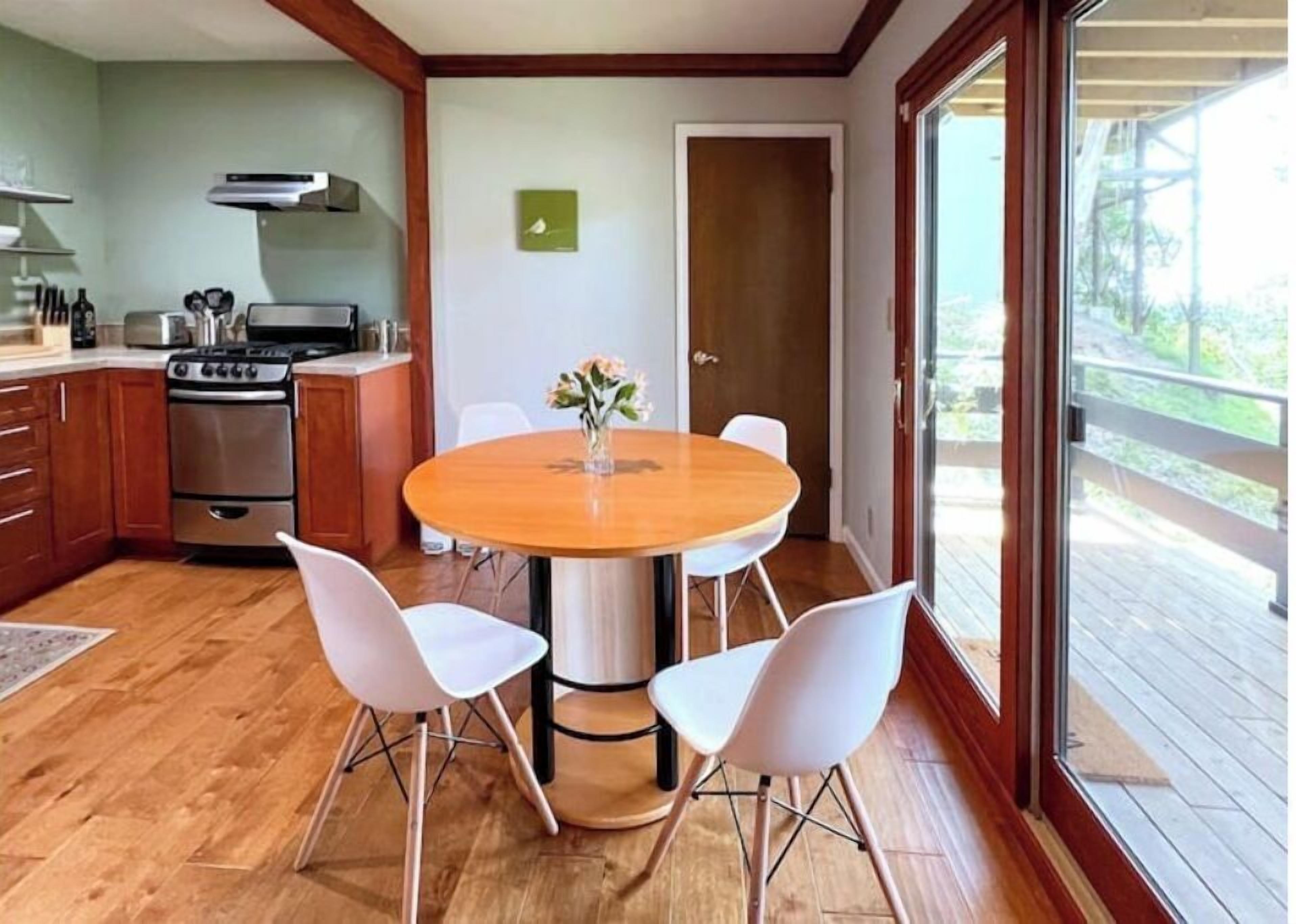 Une cuisine avec une table en bois et des chaises blanches, un poêle et une fenêtre donnant sur une terrasse.