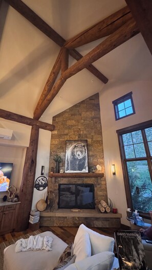 A cozy living room with a stone fireplace, wooden beams, a television, and a window with a view. - Modern Chalet on the River. Hot Tub, Fireplace , Newly furnished Fishing, Hiking (Eagle)