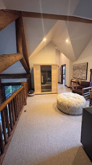 Um quarto amplo no estilo loft, com teto alto, vigas de madeira aparentes, um guarda-roupa grande, um banquinho branco fofinho e vista pela janela.