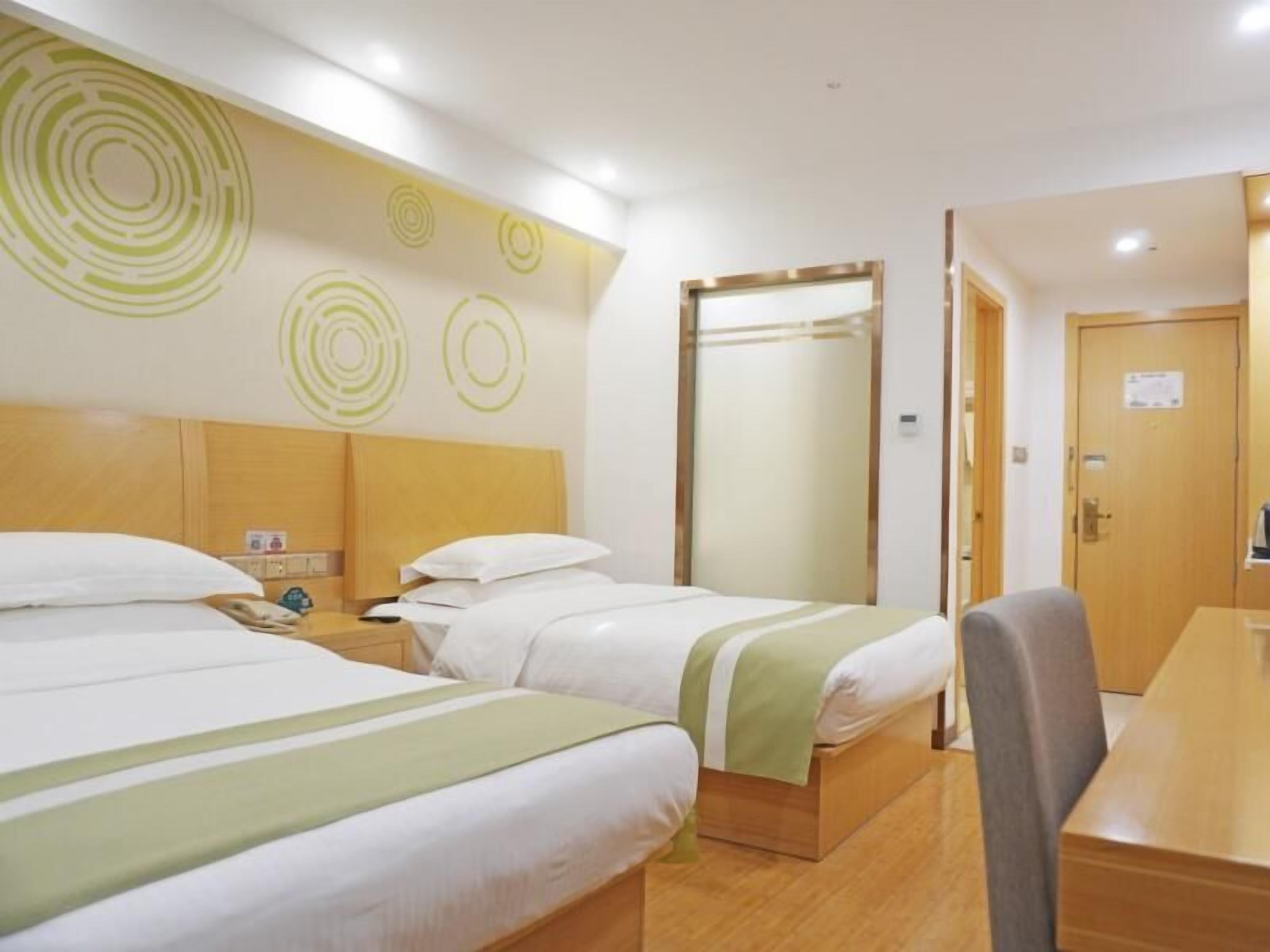Une chambre d’hôtel avec deux lits, une tête de lit en bois, un bureau et une chaise. Il y a un miroir au mur et une porte avec une pancarte.