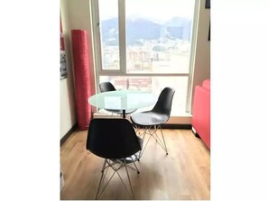 Un coin repas avec une table en verre et deux chaises, un tapis rouge et une vue sur le paysage urbain à travers de grandes fenêtres.