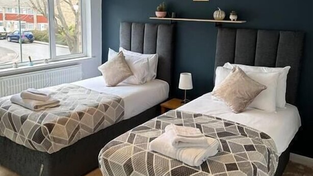 A hotel room with two beds, a window with a view of the street, and a shelf with decorative items. - Charming 2-BR House offering WIFI, Parking and a Garden | On Site Stays Welling (Welling)
