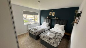 A bedroom with two beds, a window with a view of the street, a radiator, and a shelf with decorative items. - Charming 2-BR House offering WIFI, Parking and a Garden | On Site Stays Welling (Welling)
