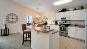 A kitchen with white cabinets, a central island with chairs, a clock on the wall, and a pendant light.
