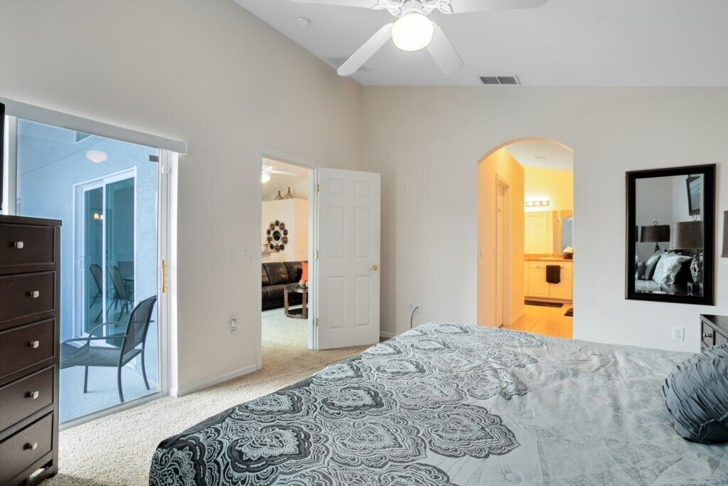 Ein Schlafzimmer mit Bett, Kommode und Deckenventilator. Eine Tür führt zu einem Badezimmer und einem Flur.