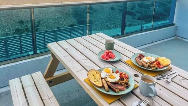 A wooden table with breakfast plates, a mug, and cutlery on a balcony overlooking a beach. - 3 bedroom accommodation in Hayling Island (Hayling Island)