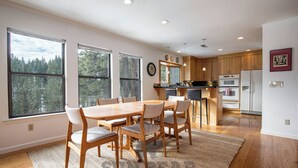 Une cuisine moderne avec des meubles en bois, un coin repas doté d’une grande table et de chaises, et de grandes fenêtres offrant une vue sur les arbres.