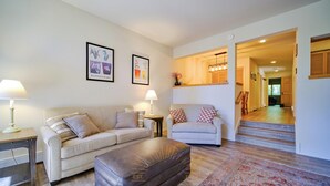 Un salon avec deux canapés, une table basse et un tapis à motifs. Des tableaux encadrés ornent le mur, et un escalier mène à une autre pièce.