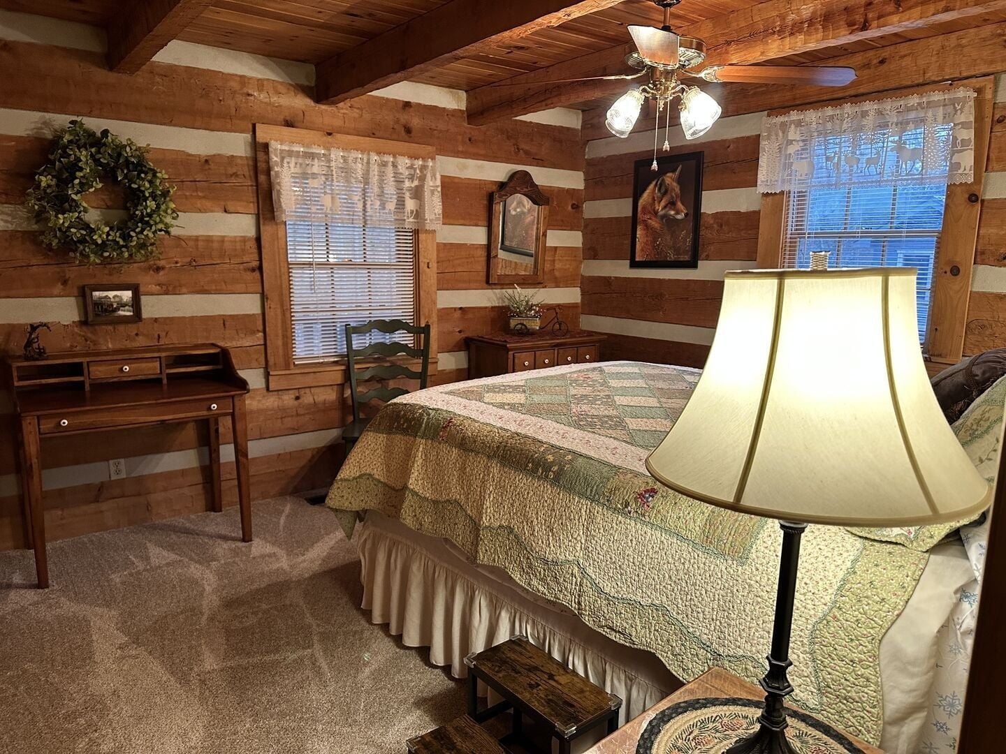 Ein gemütliches Schlafzimmer in einer Holzhütte mit einem Bett, einem Schreibtisch, einem Sessel, einer Lampe und einem Fenster mit Jalousien.