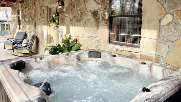 Ein Wellnessbereich mit Steinsäulen, einem Whirlpool, einem Korbsessel und einem Fenster mit Blick auf Bäume.