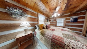 A cozy cabin-style bedroom with a wooden ceiling, a bed with a floral quilt, a nightstand with a lamp, and a wall-mounted picture.