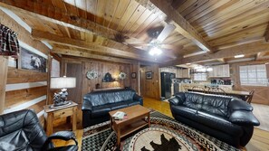 A rustic cabin interior with wooden walls, a black leather sofa, a dining table, and a kitchen area.