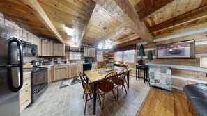 A rustic cabin interior with wooden walls, a kitchen with wooden cabinets, a dining area with a table and chairs, and a fireplace.