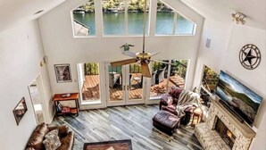 Un salon spacieux avec un haut plafond, une cheminée en pierre, un grand téléviseur et une vue sur un lac, visible à travers de grandes fenêtres.