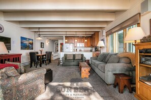 A living room with a sofa, a dining table, and chairs, a kitchen with wooden cabinets, and a large window. - Betty's Place - Beachfront w Lake Access (LA99958) (Incline Village)