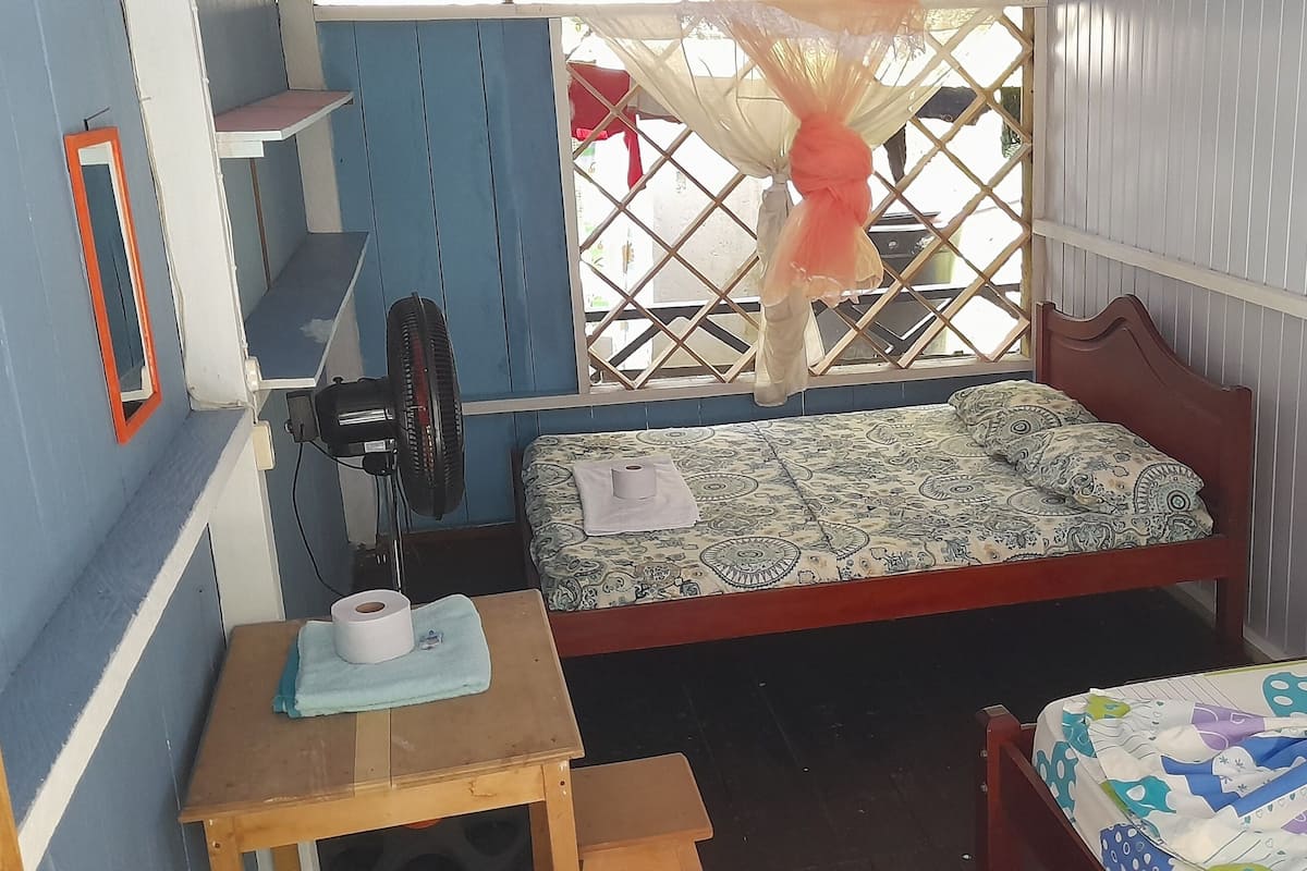 A small, compact room with a single bed, a small table with a towel and toilet paper, a fan, and a window with a mosquito net.