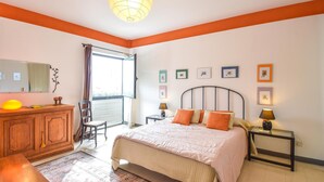 Une chambre à coucher comprenant un lit, des tables de chevet, une commode, une chaise, une fenêtre et des tableaux accrochés au mur.