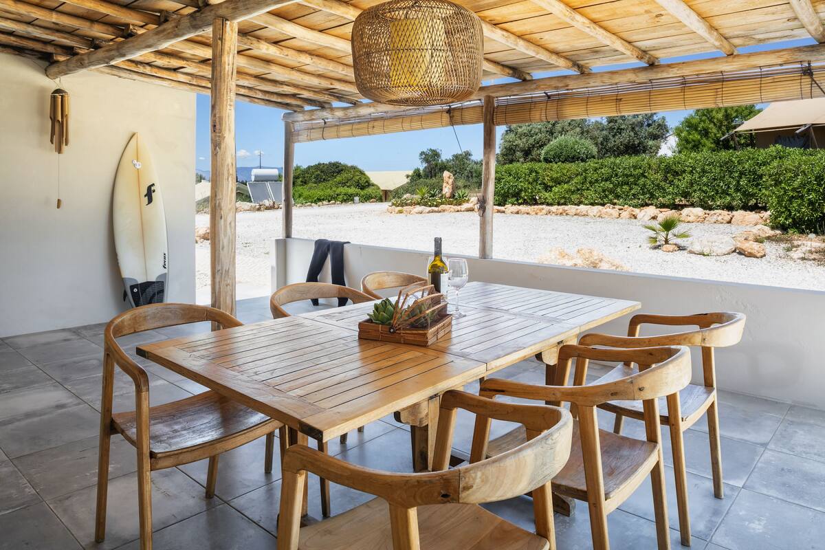 A wooden table with six chairs, a surfboard, and a bottle of wine on a patio with a view of the beach and greenery.