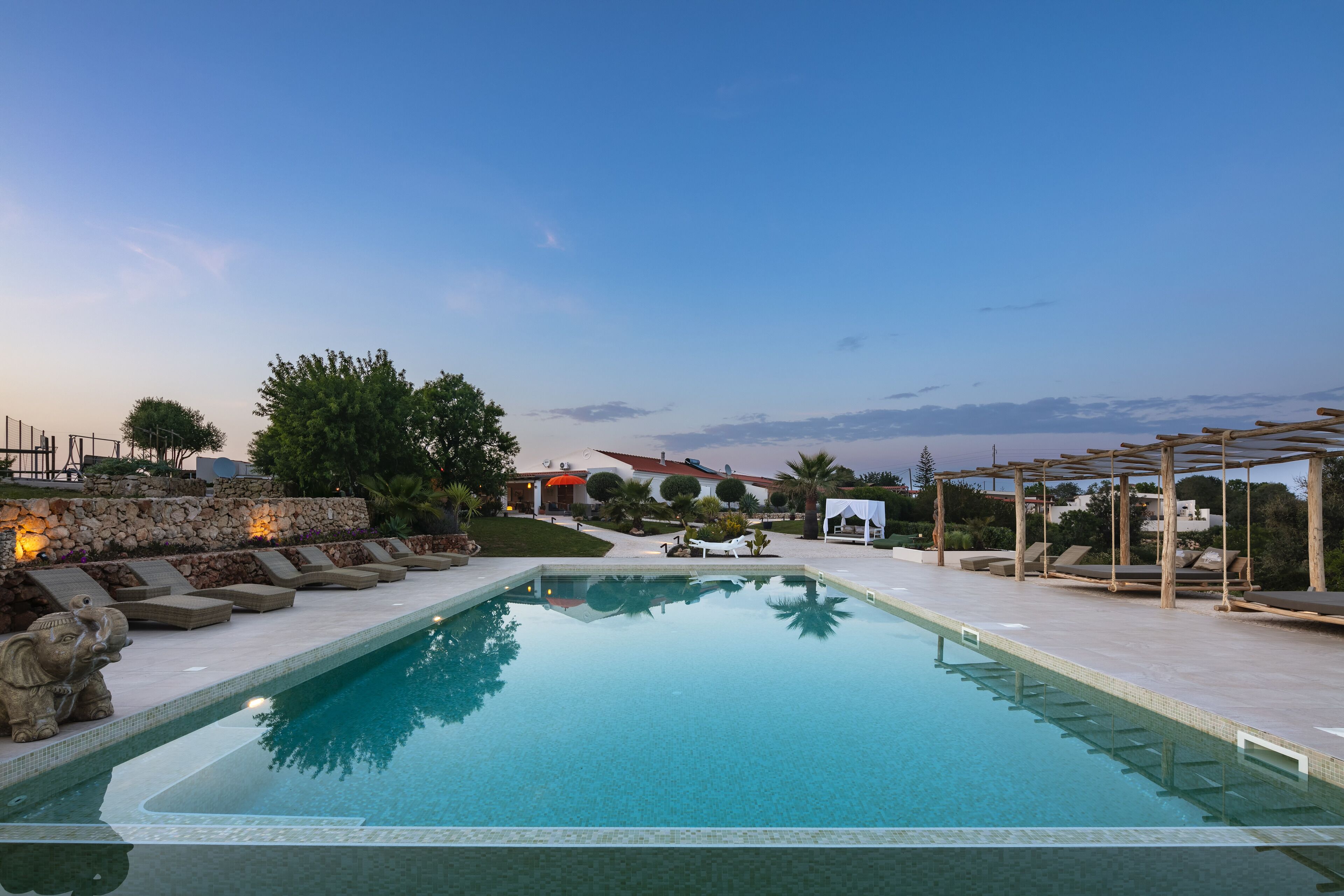 A rectangular swimming pool with clear blue water, surrounded by lounge chairs and a stone wall.
