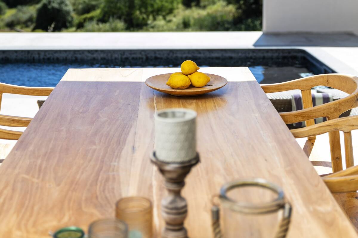 A wooden dining table with a bowl of lemons, a candle, and glasses, overlooking a swimming pool and lush greenery.
