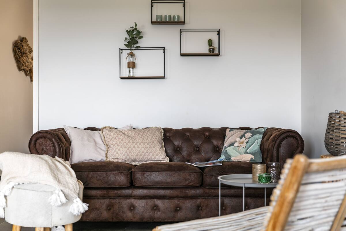A brown tufted sofa with patterned cushions, a white chair, and a small round table with decorative items.