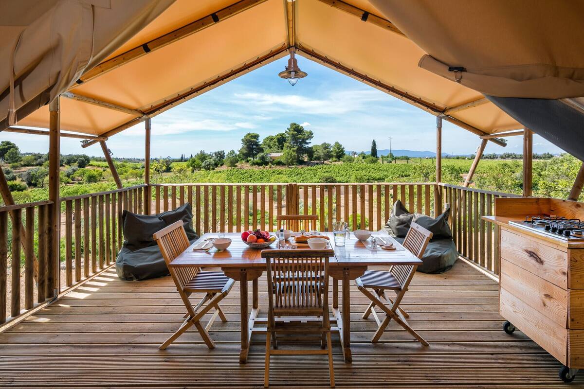 A wooden deck with a table set for dining, surrounded by cushioned chairs, under a canopy.