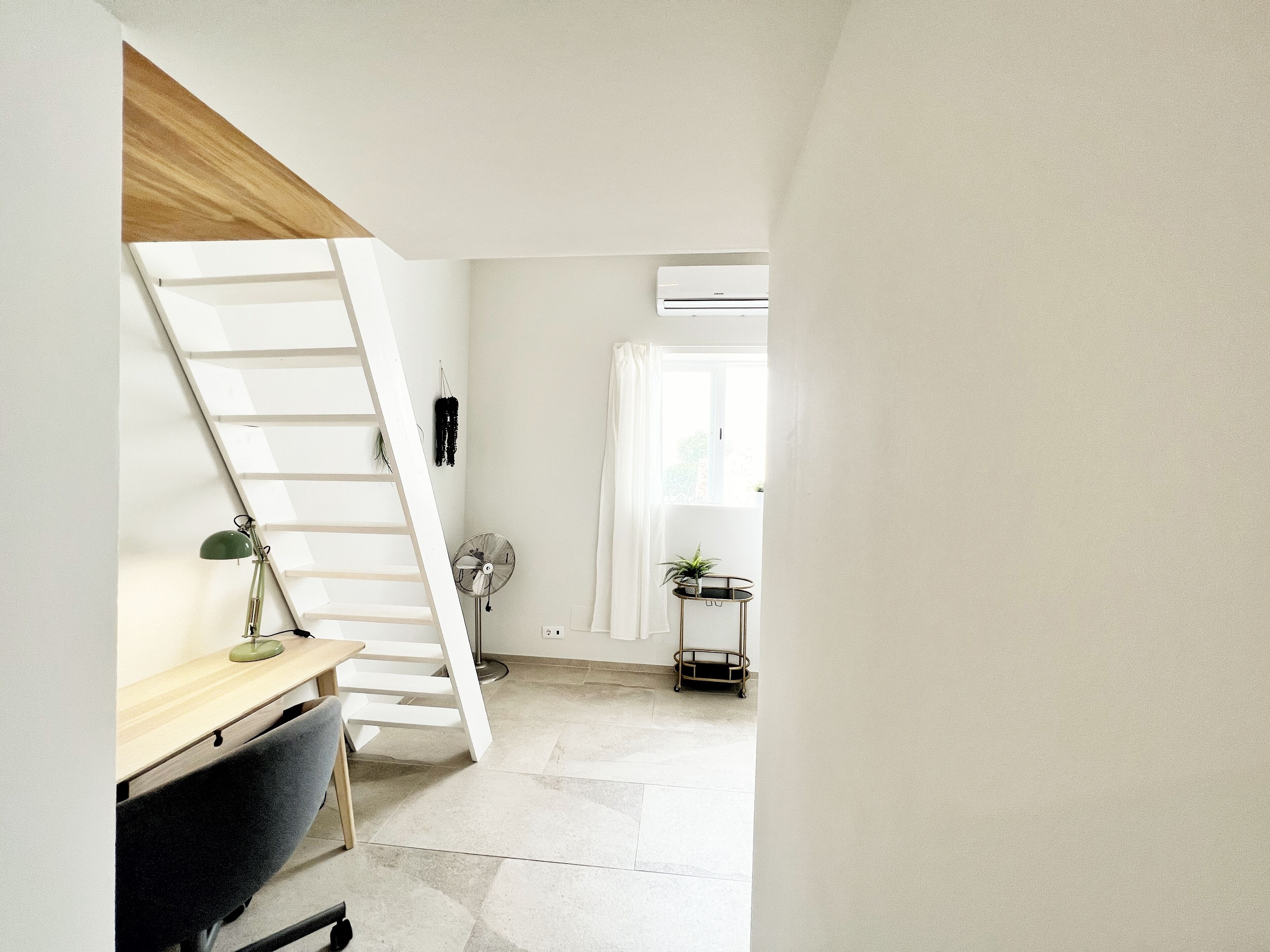 A room with a desk, a chair, a ladder, a fan, a small table with a plant, and an air conditioning unit.
