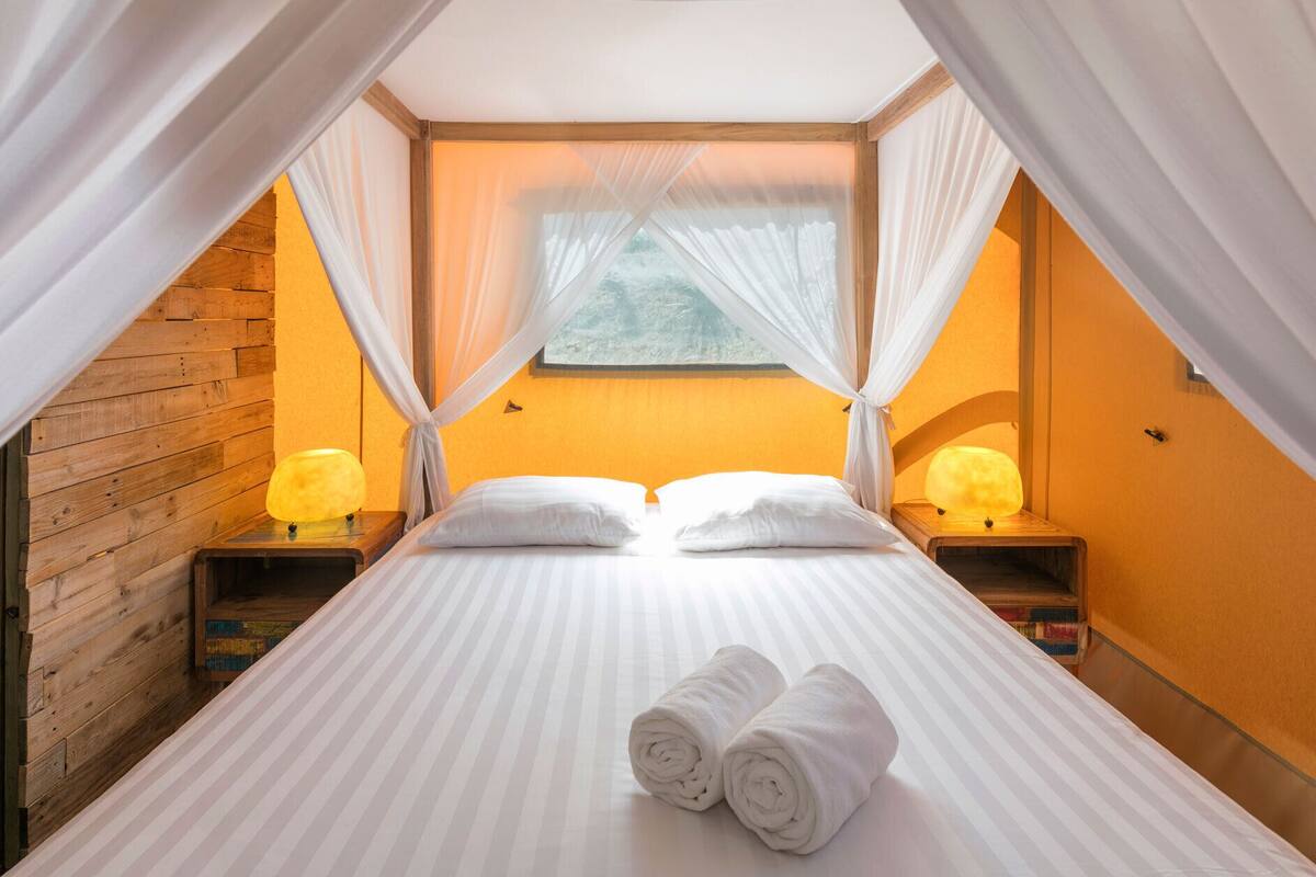 A bedroom with a bed, white curtains, two bedside lamps, and a window with a view.