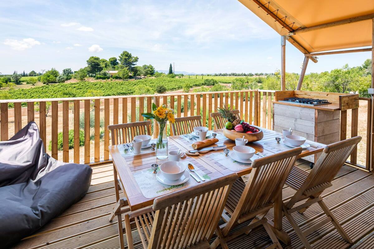 An outdoor dining area with a table set for two, a wooden deck, and a view of vineyards in the background.
