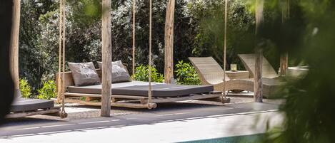 A pool area with a wooden swing chair, a lounge chair, and a cushioned bench.