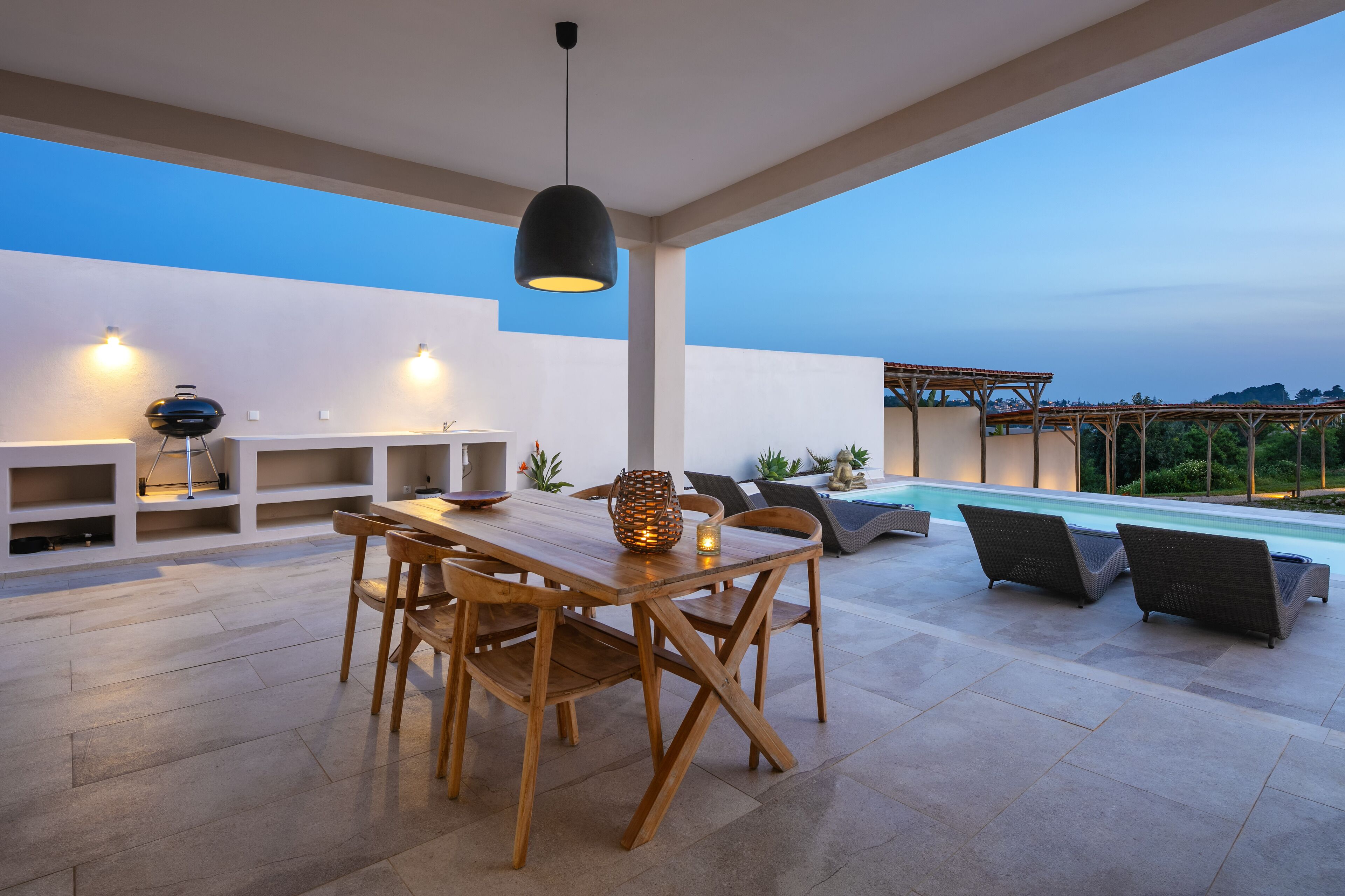 An outdoor dining area with a wooden table and chairs, a hanging light fixture, and a pool in the background.
