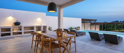 An outdoor dining area with a wooden table and chairs, a hanging light fixture, and a pool in the background.