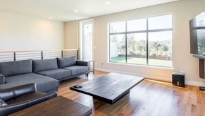 En moderne stue med en grå hjørnesofa, et sofabord og et stort vindue med udsigt over grønne områder.