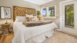 A bedroom with a large bed, a wooden headboard, a mirror, a lamp, and a view of greenery through the window. - Misty Mountain | Views, Hot Tub, Fire Pit + Biltmore Pass Near Asheville (Asheville)