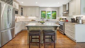 A modern kitchen with stainless steel appliances, a large island with stools, and a view of greenery through the windows. - Misty Mountain | Views, Hot Tub, Fire Pit + Biltmore Pass Near Asheville (Asheville)