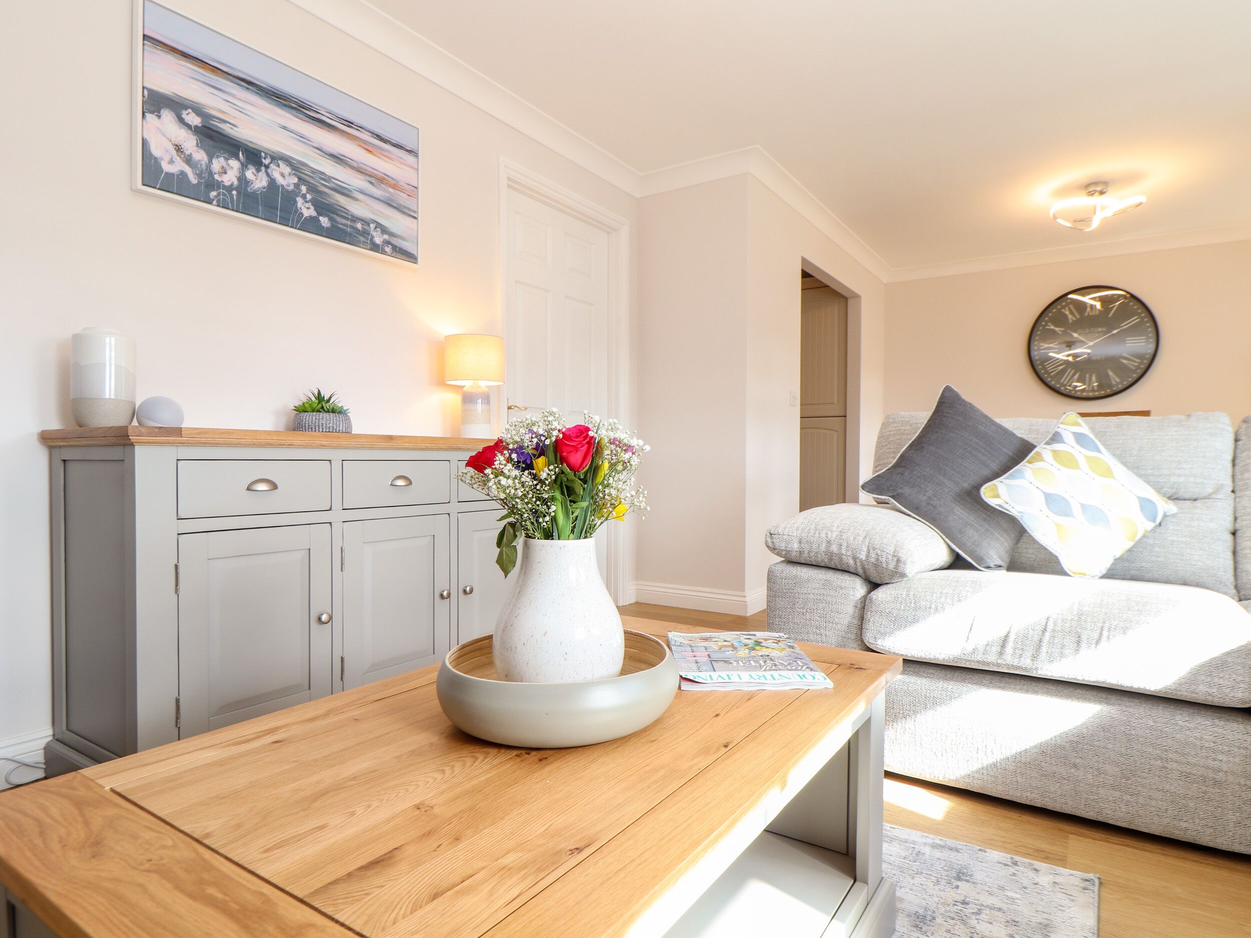 A modern living room with a wooden coffee table, a white vase with flowers, a grey sofa with patterned cushions, and a wall clock.