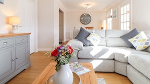 Una sala de estar con un sofá gris, una mesa de centro de madera, un jarrón blanco con flores y un reloj en la pared.